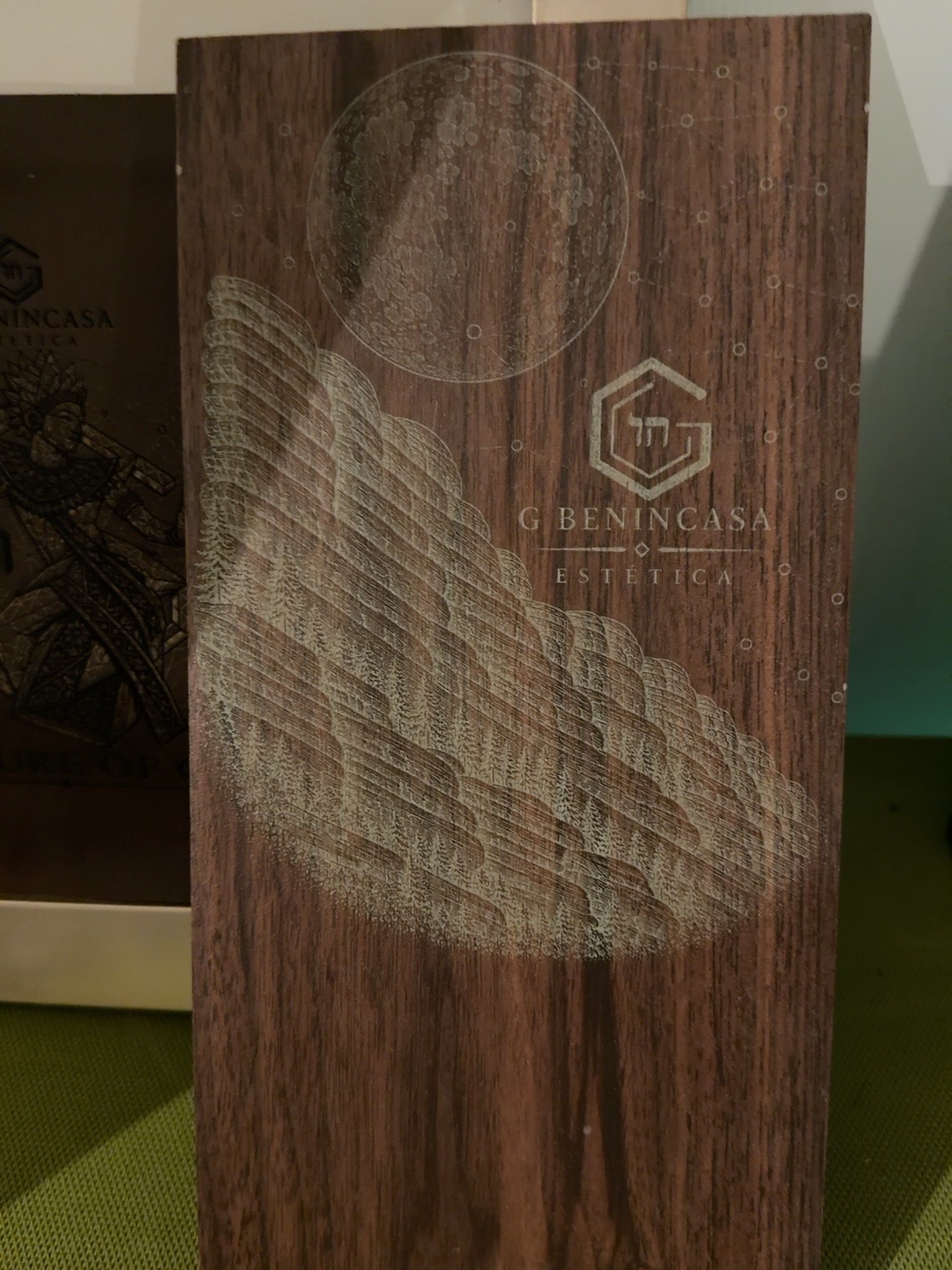 Existing photograph of a wood-look porcelain panel with a pale engraved wing, moon, orbital dots and G Benincasa lettering.